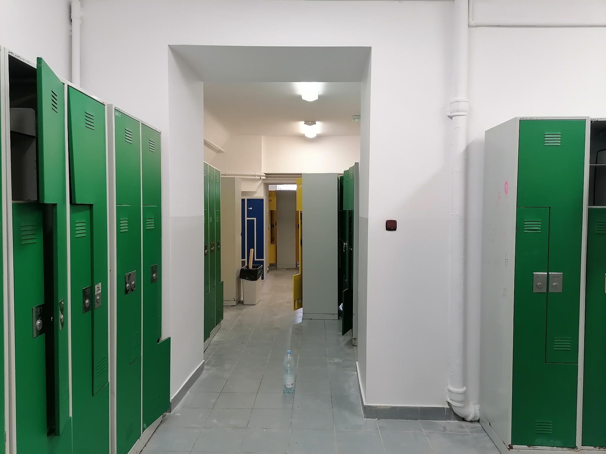 Korytarz z rzędem zielonych szafek, niektóre z otwartymi drzwiczkami, białe ściany i szara podłoga z butelką wody w centrum kadru.