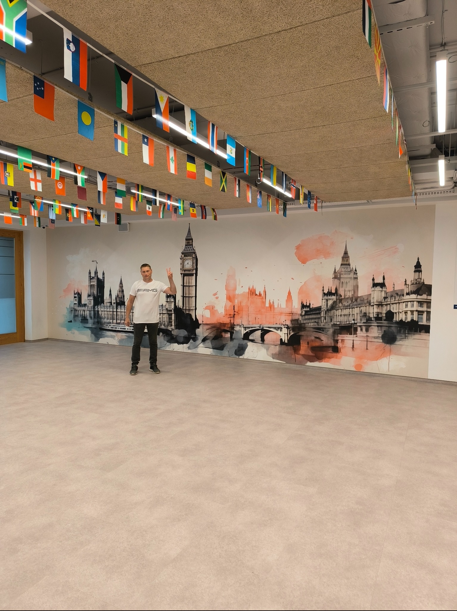 Wnętrze biura z dekoracyjną malowaną ścianą przedstawiającą panoramę Londynu, ozdobione flagami różnych państw pod sufitem, z mężczyzną w koszulce stojącym na tle ściany.