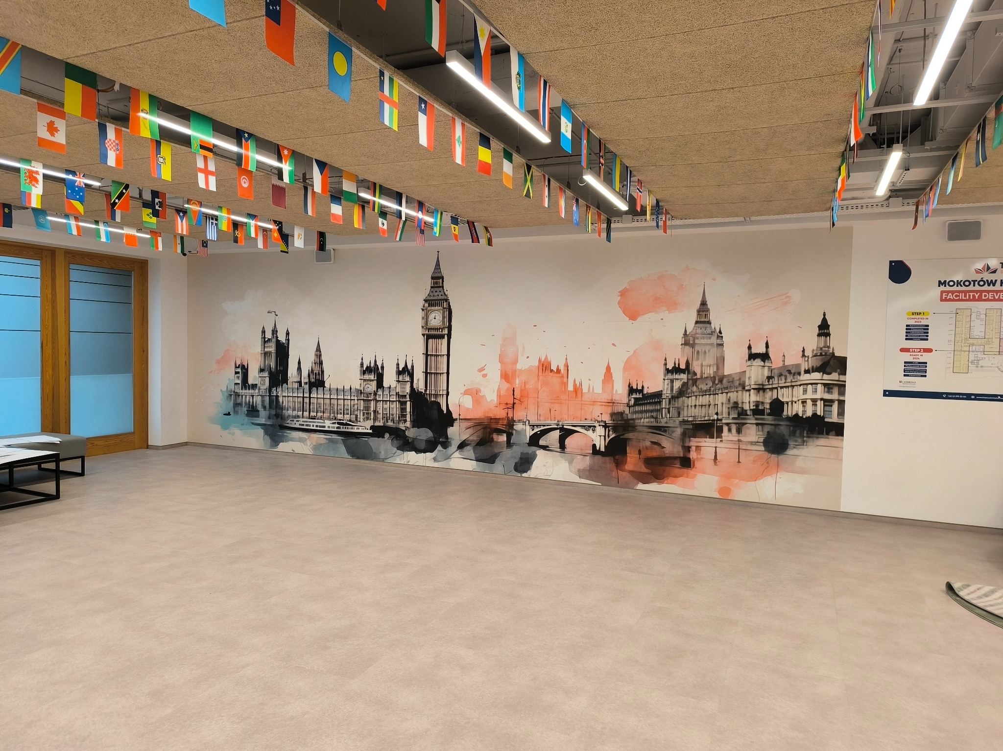Duża, pusta przestrzeń biurowa z muralem przedstawiającym panoramę Londynu w odcieniach szarości i pomarańczy, udekorowana flagami różnych państw podwieszonymi pod sufitem.