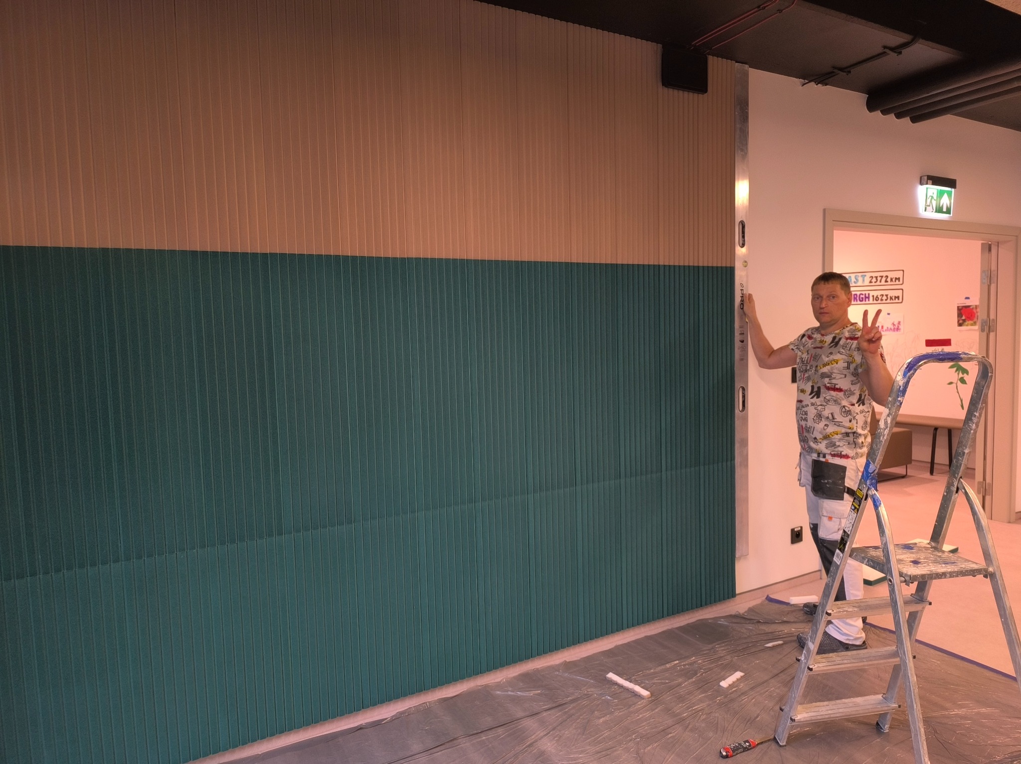 Ściana wykończona panelami w dwóch odcieniach, zielonym i beżowym, z widocznym pracownikiem sprawdzającym poziom pionu za pomocą poziomicy, drabina malarska w pomieszczeniu biurowym.