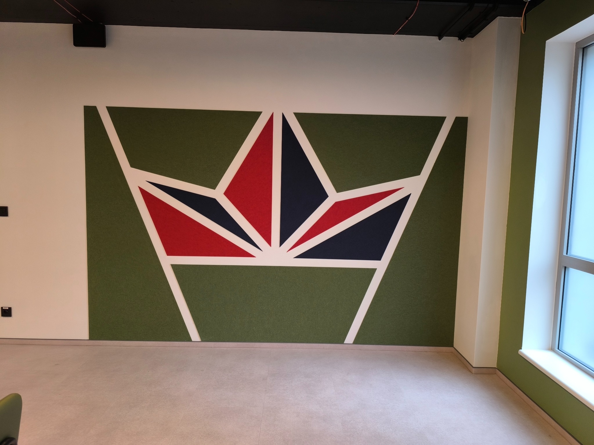Geometryczny wzór w kształcie korony z czerwonymi i granatowymi elementami na zielonym tle, otoczony białymi liniami na białej ścianie w biurze.