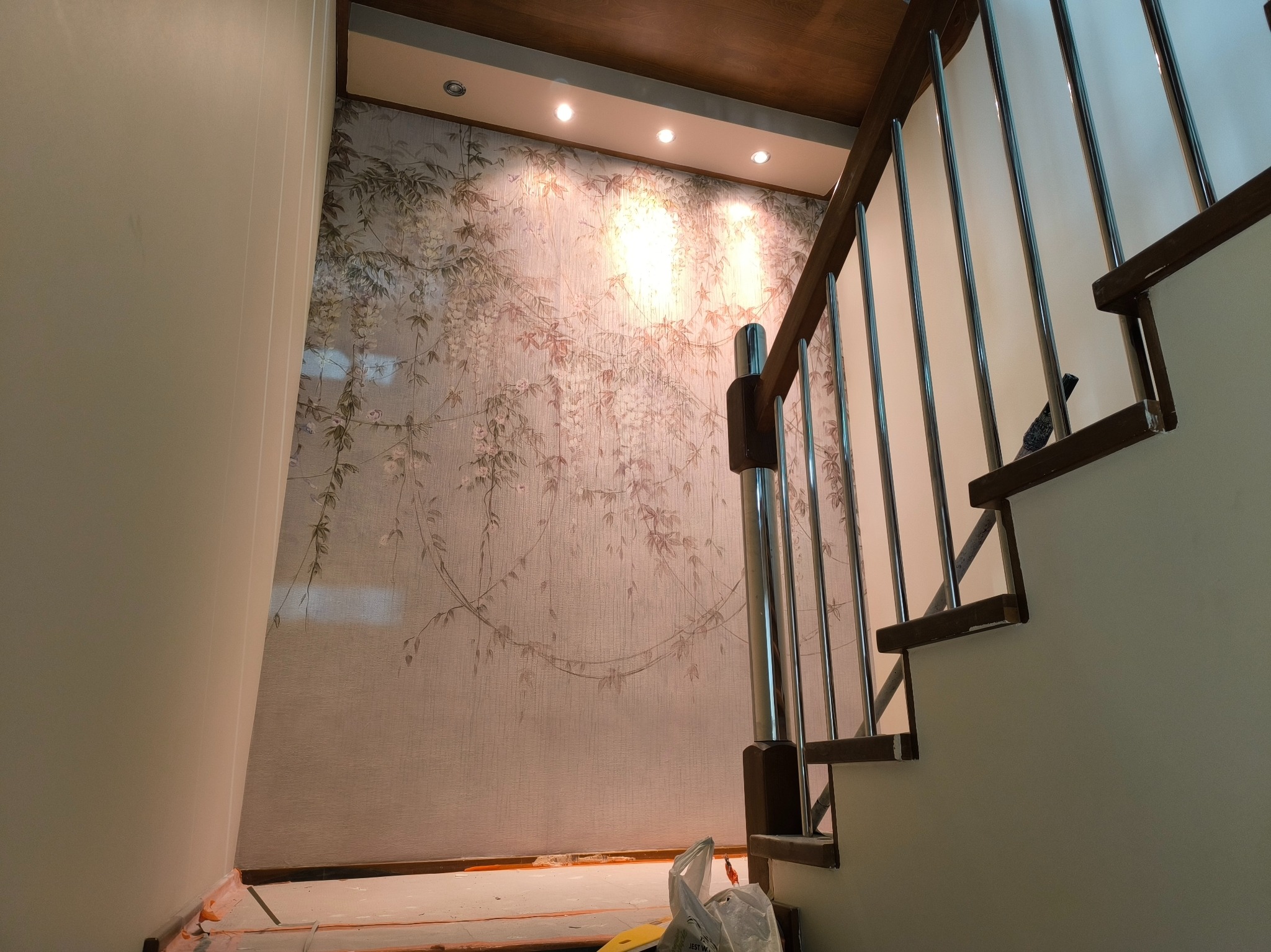 Ściana z tapetą w motywy roślinne, oświetlona punktowo, widoczna przy schodach z metalową balustradą i drewnianymi elementami.