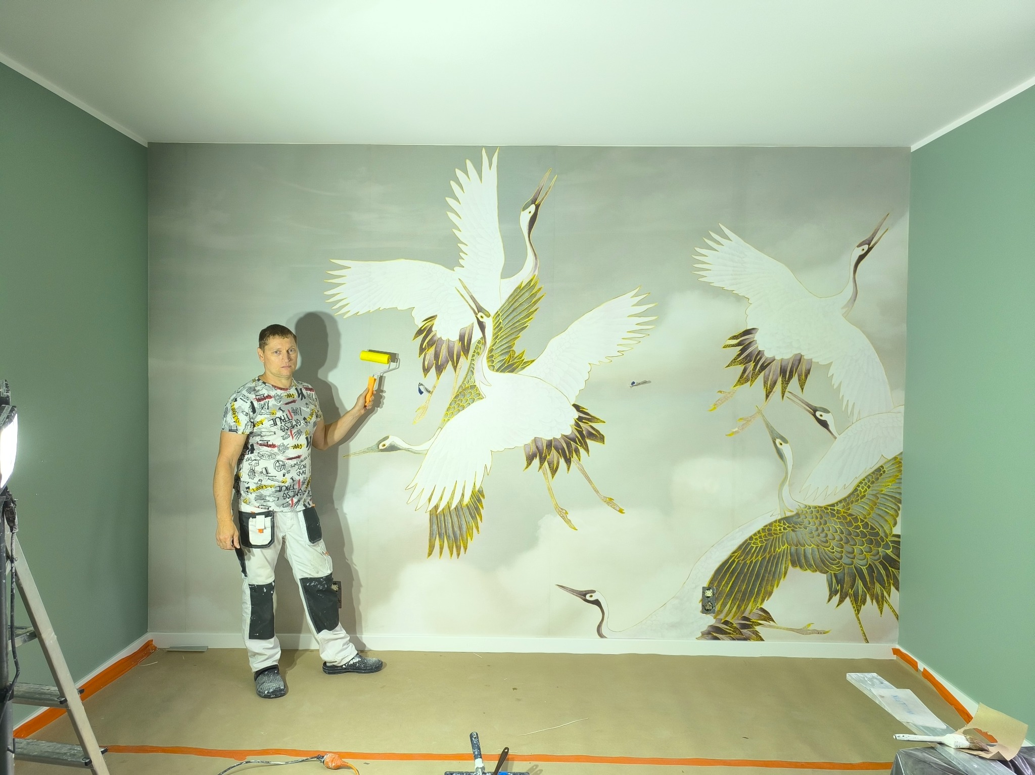 Pokój po remoncie z pomalowaną ścianą z motywem żurawi, malarz trzyma wałek do malowania, widoczne narzędzia i akcesoria malarskie.