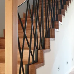 Drewniane schody z oryginalną, geometryczną balustradą z czarnych prętów, widoczne detale wykończenia i fragment podłogi z folią ochronną.