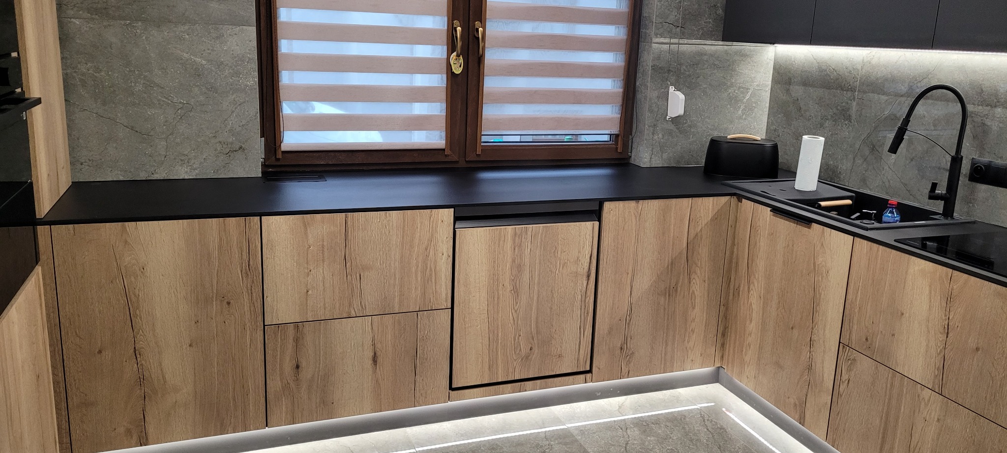 Nowoczesna kuchnia z drewnianymi frontami szafek, czarnym blatem i szarymi płytkami na ścianie, oświetlona listwą LED pod szafkami i punktowym oświetleniem nad blatem.