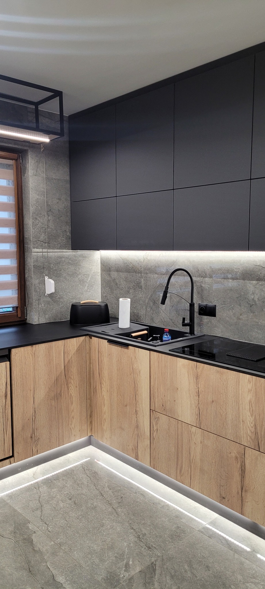Nowoczesna kuchnia z drewnianymi frontami szafek dolnych i grafitowymi szafkami górnymi, czarnym blatem, szarą mozaiką na ścianie, oświetleniem LED pod szafkami i na podłodze, czarną armaturą...