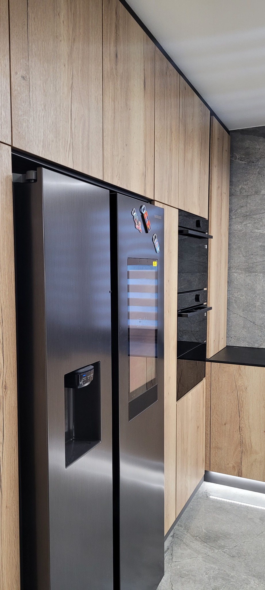 Nowoczesna kuchnia z drewnianymi frontami szafek, czarnym wykończeniem i dużą, dwudrzwiową lodówką Samsung z wyświetlaczem i kostkarką do lodu.