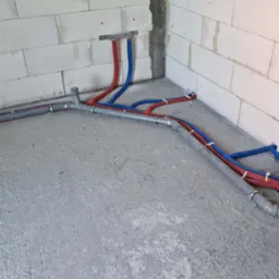 Surowy stan: Instalacja rur grzewczych (czerwone i niebieskie) i kanalizacyjnych (szare) w narożniku pomieszczenia z bloków betonowych, widoczne mocowania i złącza.