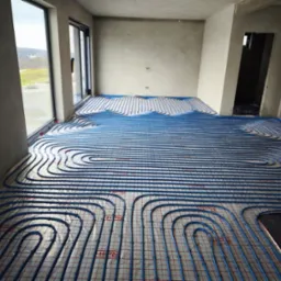 Niebieskie rury instalacji ogrzewania podłogowego ułożone na izolacji z siatką w surowym wnętrzu, widok z góry na instalację przed wylaniem posadzki.