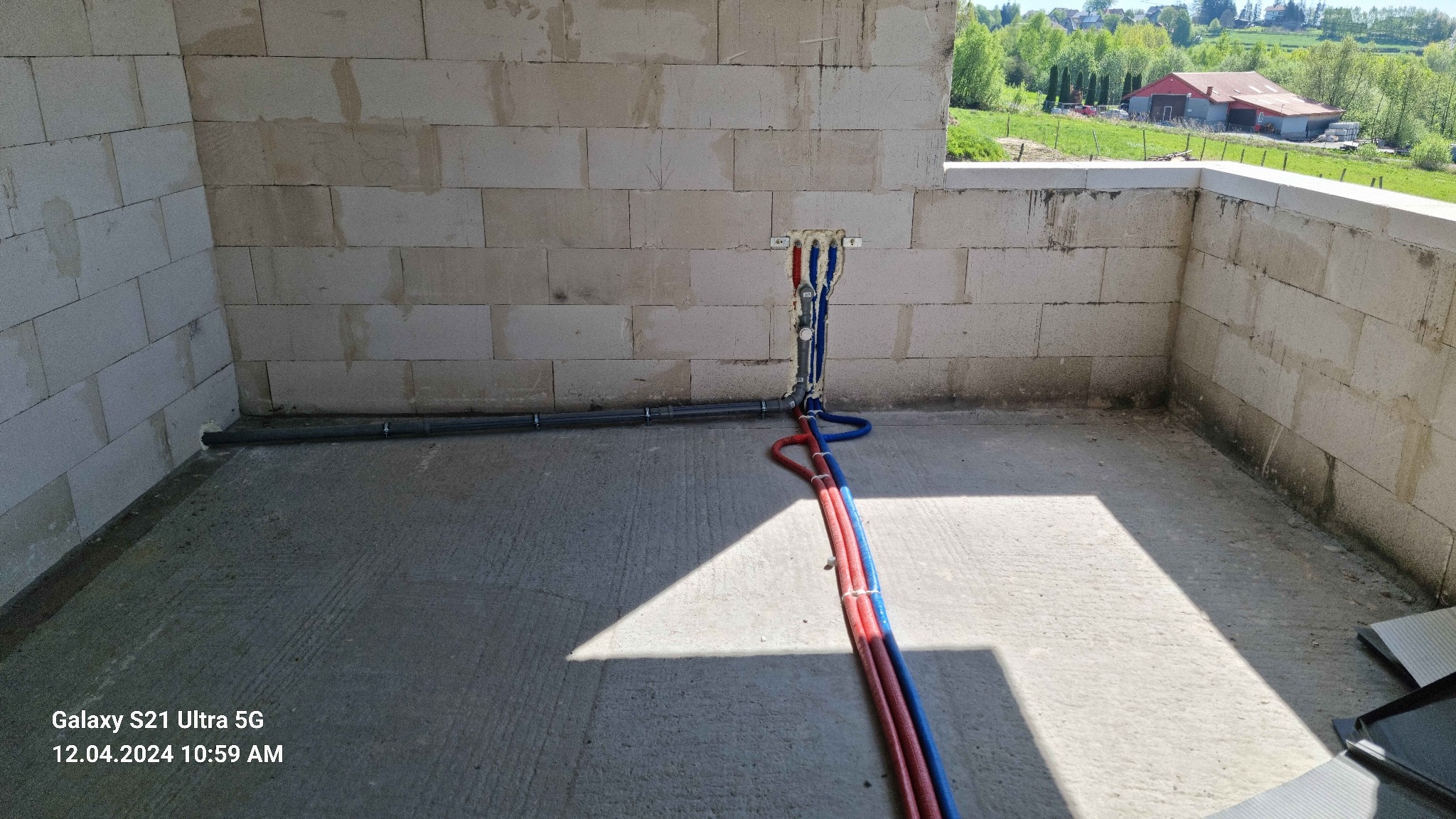 Surowe ściany z betonu komórkowego w trakcie budowy, z wyprowadzonymi rurami instalacji wodnej (czerwone i niebieskie) i szarą rurą kanalizacyjną, widok z góry na podłogę w cieniu i na zewnątrz...