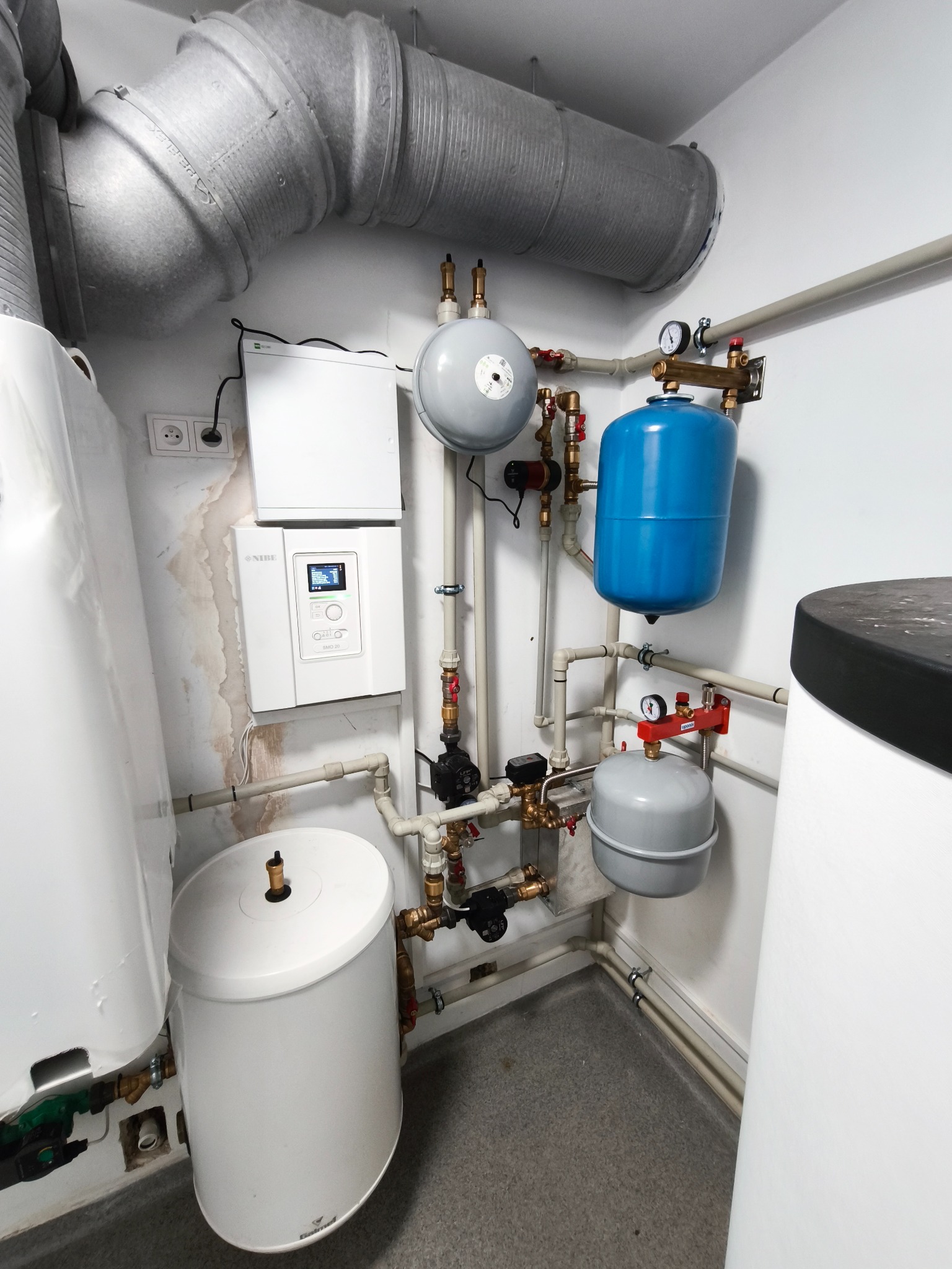 Kompaktowa kotłownia z piecem, zbiornikami wyrównawczymi i orurowaniem, widok z góry na szare płytki podłogowe i białe ściany z widocznymi śladami wilgoci.