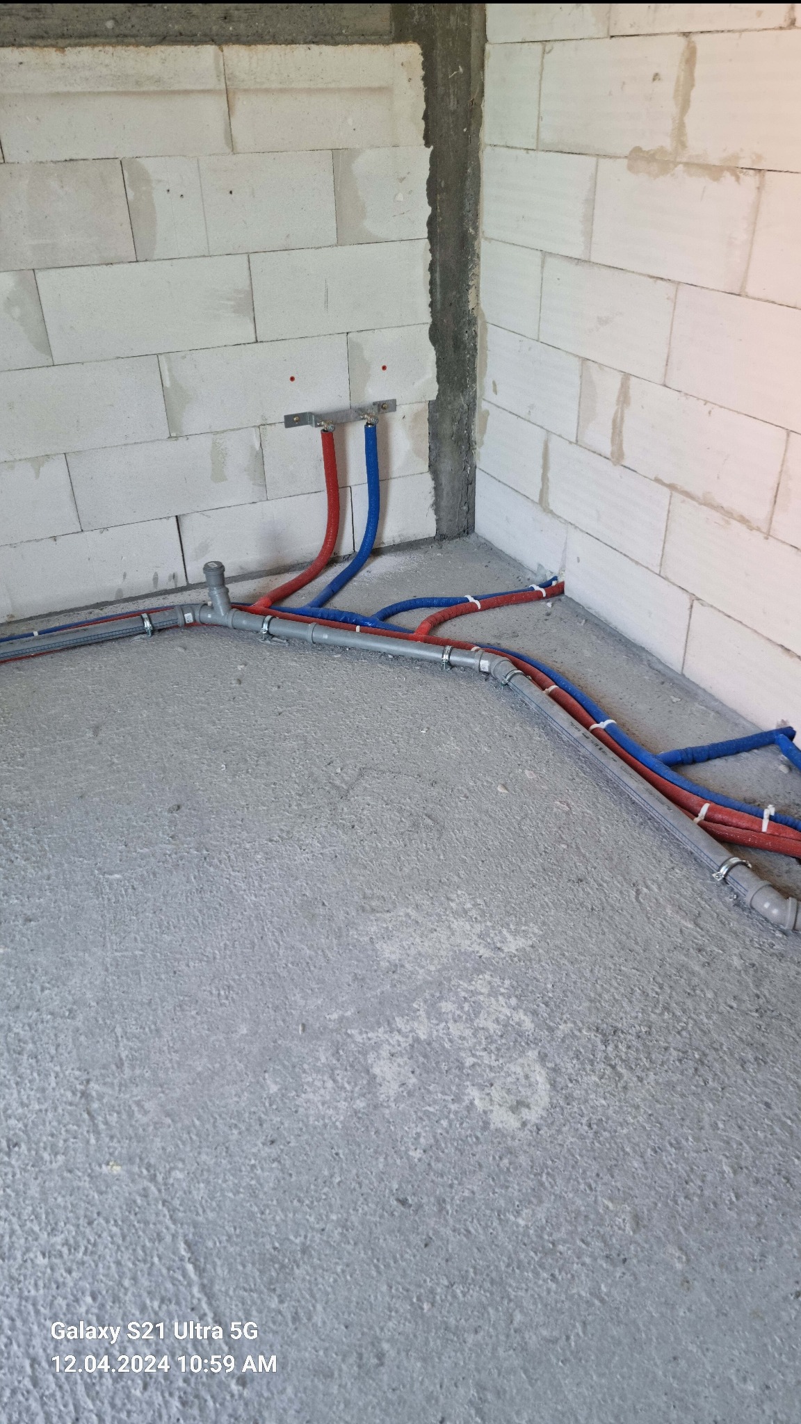 Surowy stan: Instalacja rur grzewczych (czerwone i niebieskie) i kanalizacyjnych (szare) w narożniku pomieszczenia z bloków betonowych, widoczne mocowania i złącza.