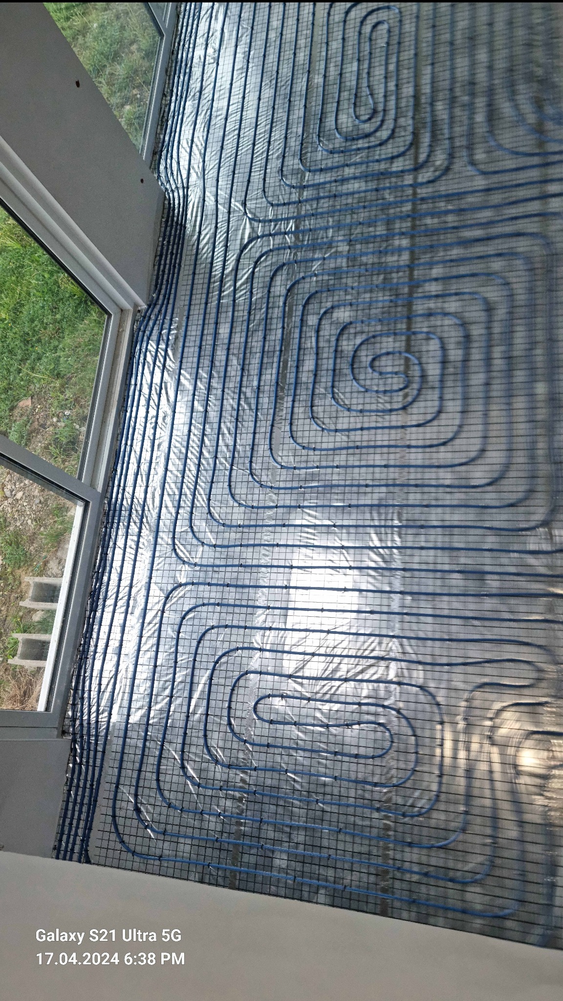 Niebieskie rurki ogrzewania podłogowego ułożone w pętle na srebrnej folii izolacyjnej, widok z góry, fragment okna z widokiem na zieleń.