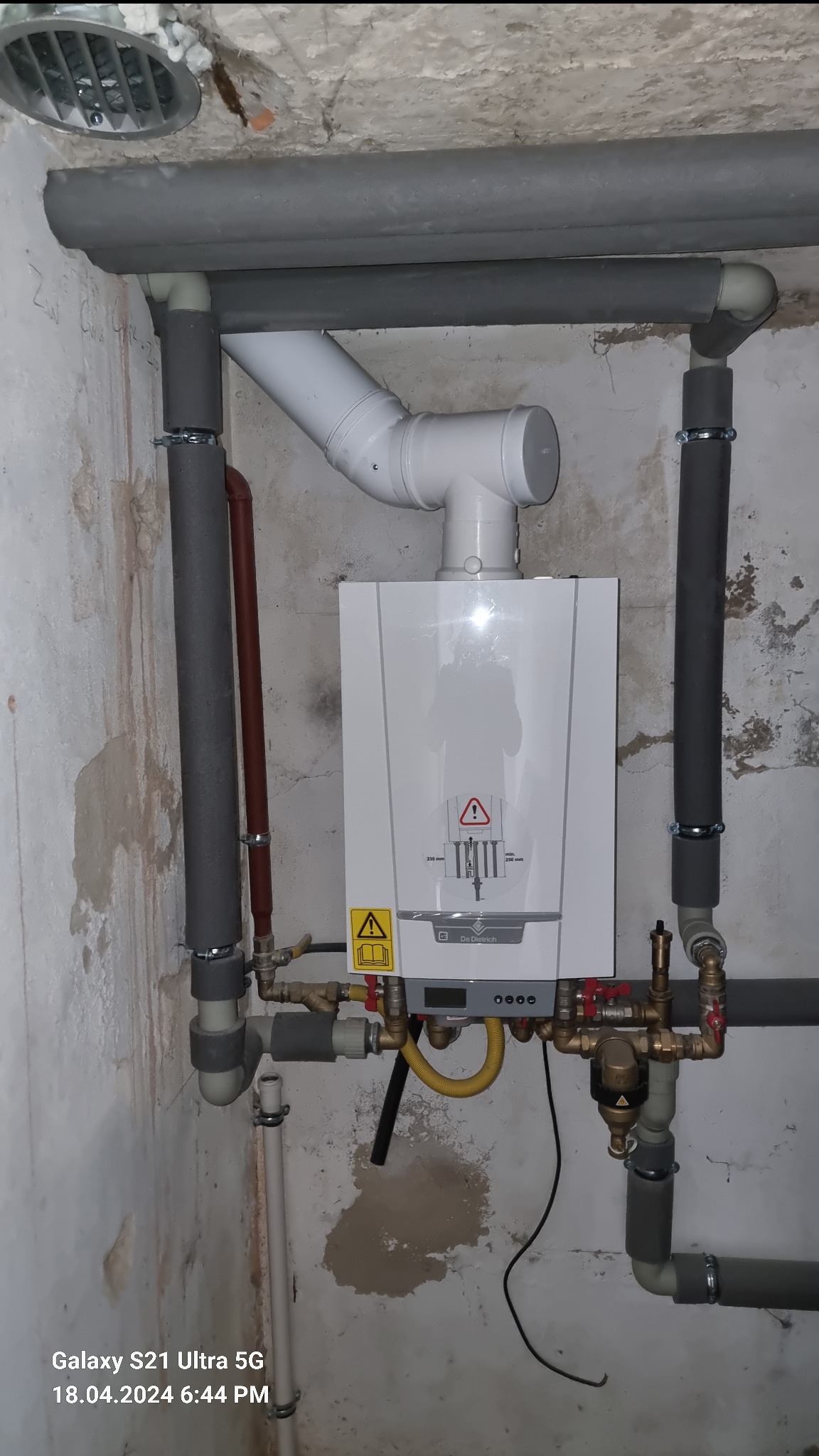 Zamontowany gazowy piec grzewczy De Dietrich z podłączonymi rurami w piwnicy z surowymi ścianami, widoczne rury izolowane szarą pianką i liczne zawory