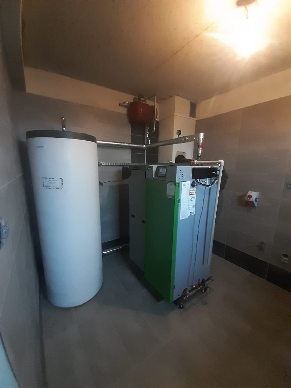 Kotłownia z piecem na paliwo stałe z zielonym panelem bocznym i białym zasobnikiem wody, widoczne rury i instalacja, szare płytki na ścianach i podłodze.