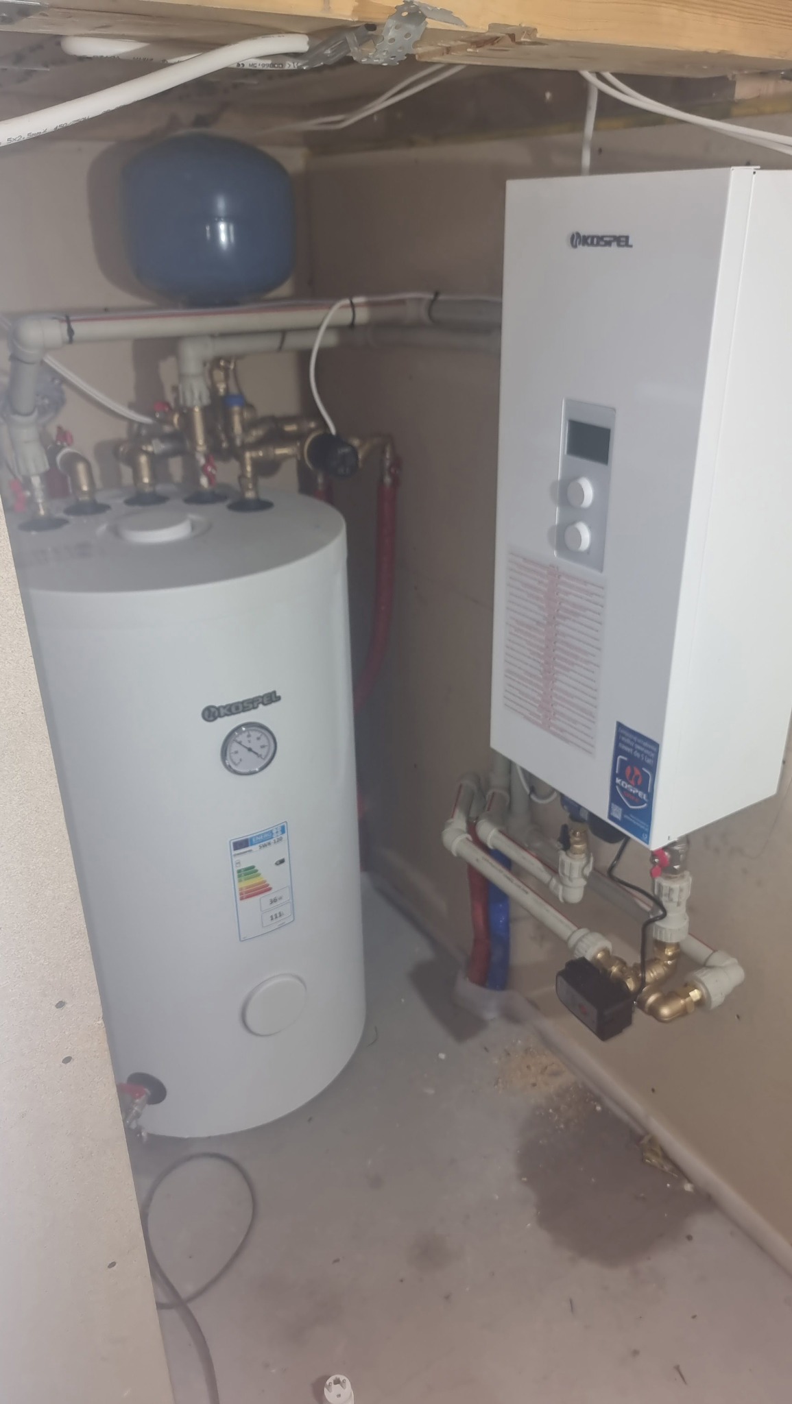 Biały, cylindryczny bojler Kospel z widocznymi rurami miedzianymi i plastikowymi, obok biały piec gazowy Kospel, zamontowane w pomieszczeniu ze ścianami w kolorze beżowym i surowym sufitem.