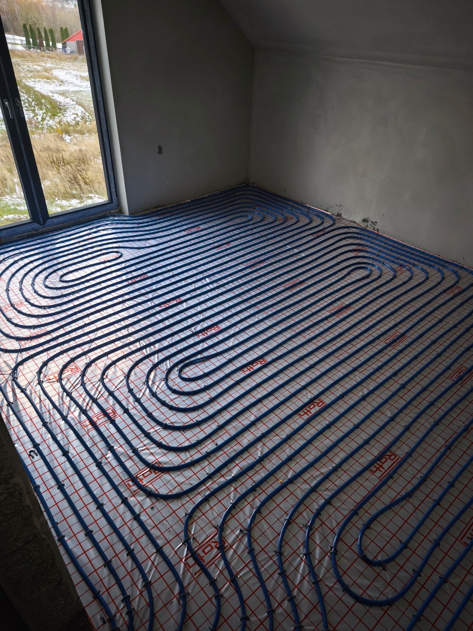 Niebieskie rury ogrzewania podłogowego ułożone spiralnie na folii z czerwoną siatką w surowym wnętrzu w Skawinie, widok z góry na instalację przed zalaniem wylewką.
