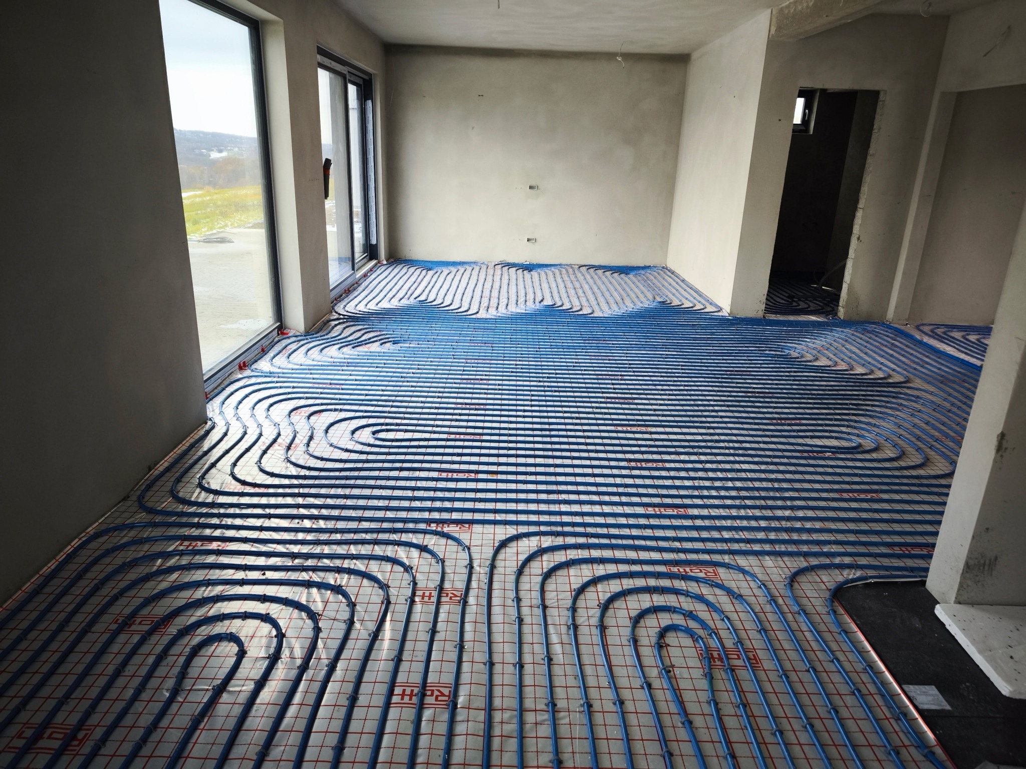 Niebieskie rury instalacji ogrzewania podłogowego ułożone na izolacji z siatką w surowym wnętrzu, widok z góry na instalację przed wylaniem posadzki.