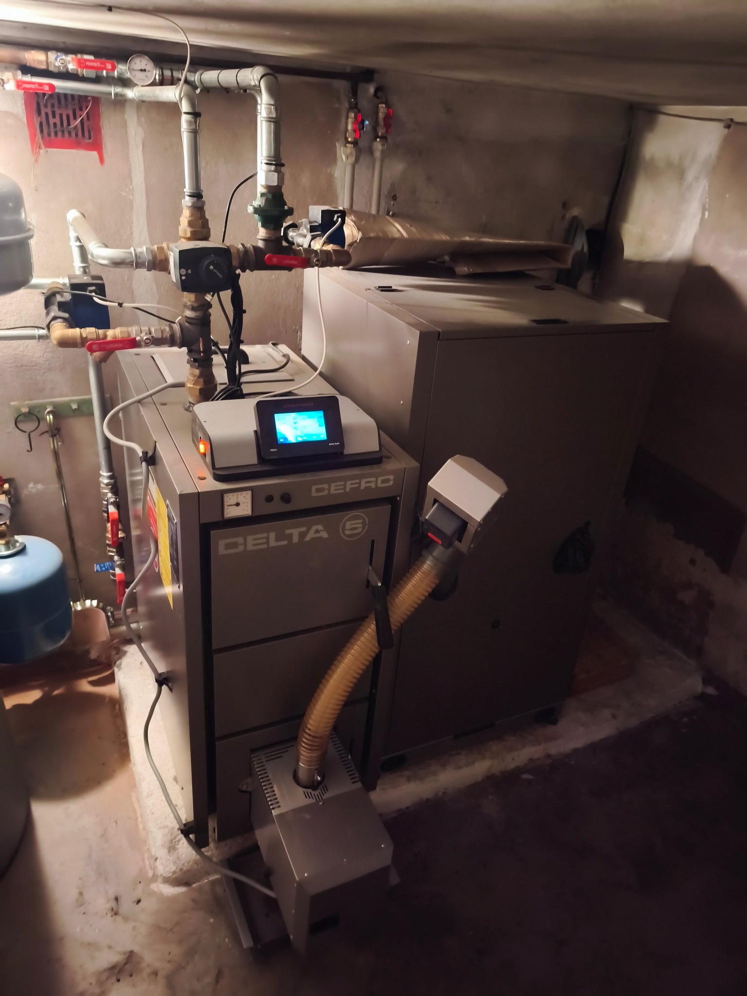 Szary piec Delta S z podajnikiem, widoczne rury instalacji grzewczej i sterownik z niebieskim wyświetlaczem w kotłowni.