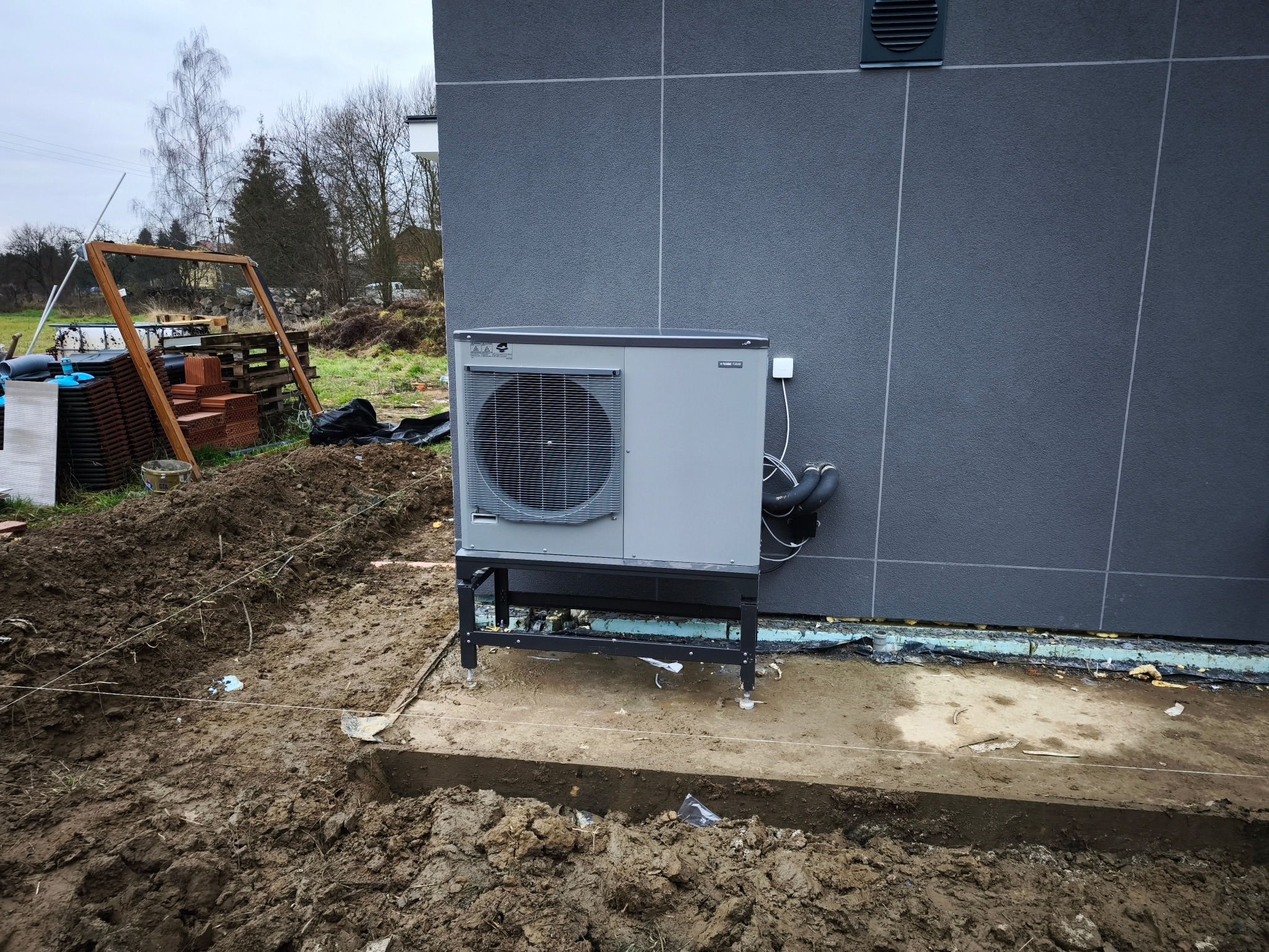 Szara pompa ciepła zamontowana na czarnym stelażu przy szarej ścianie budynku w trakcie prac ziemnych, widoczne wykopane fundamenty.