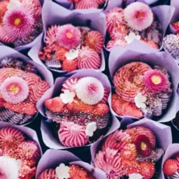 Różowe babeczki w liliowych papilotkach zdobione kremem, kwiatami i posypką, tworzące słodki bukiet. Widok z góry prezentuje bogactwo detali i artystyczne wykończenie.