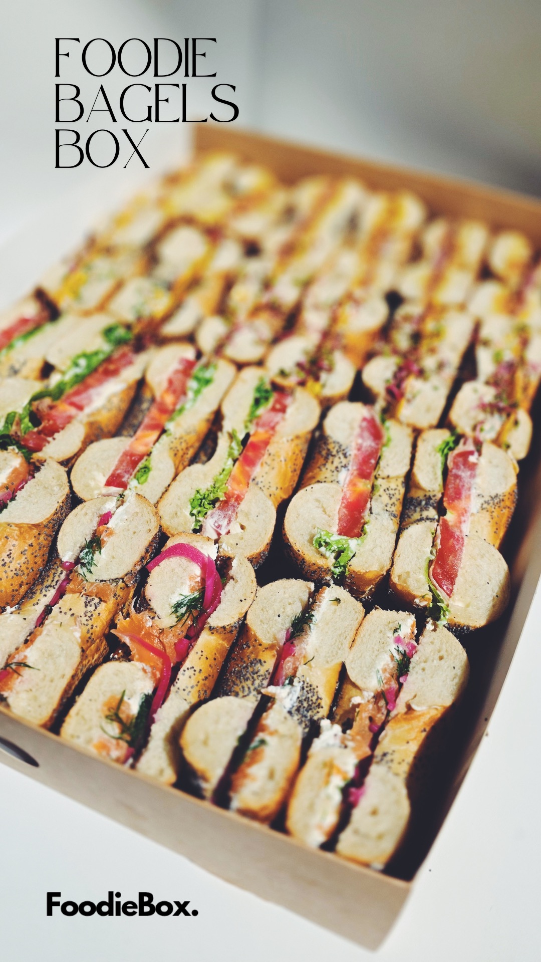 Kartonowe pudełko pełne przekrojonych bajgli z różnymi dodatkami: łososiem, warzywami i kremowym serkiem. Widoczne logo 'Foodie Bagels Box'.