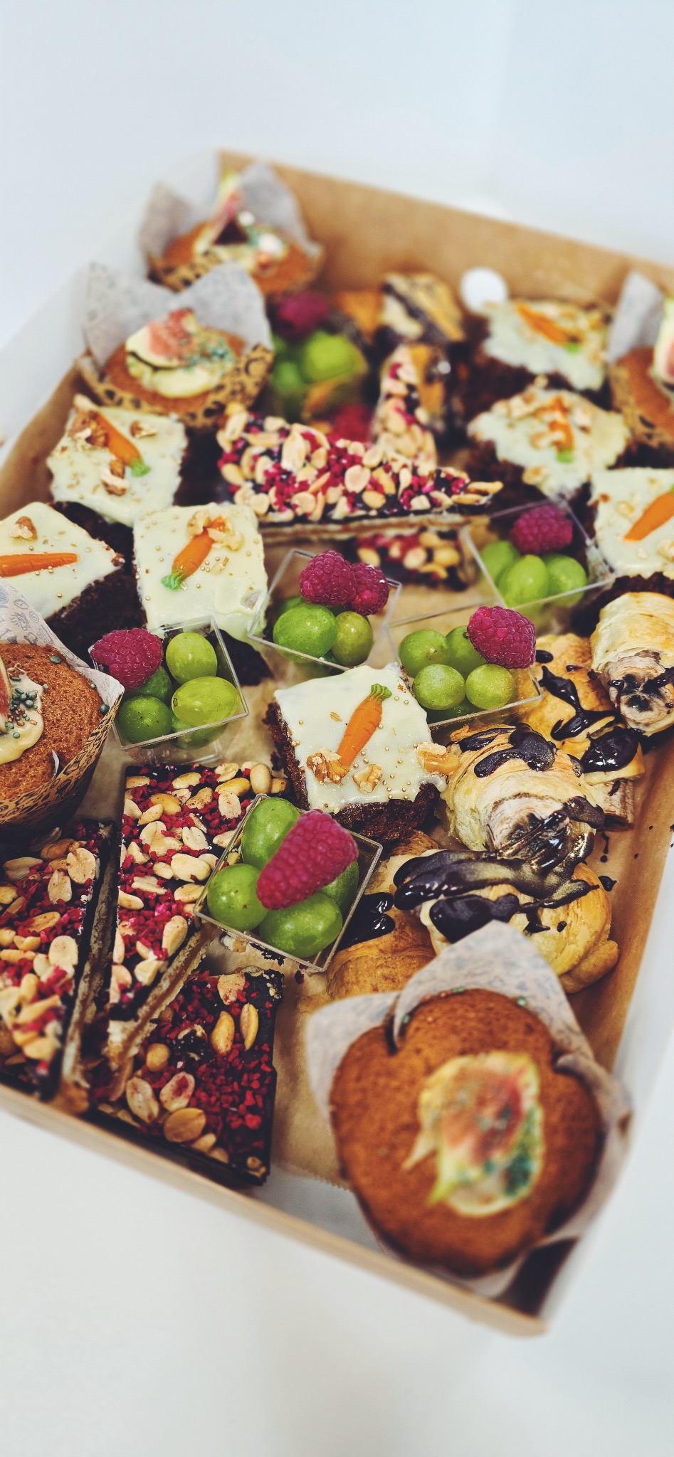 Różnorodne, apetyczne przekąski w kartonowym pudełku: babeczki z figami, ciastka z marchewką, batoniki z orzechami i malinami, owoce w małych pojemnikach, rogaliki z czekoladą.