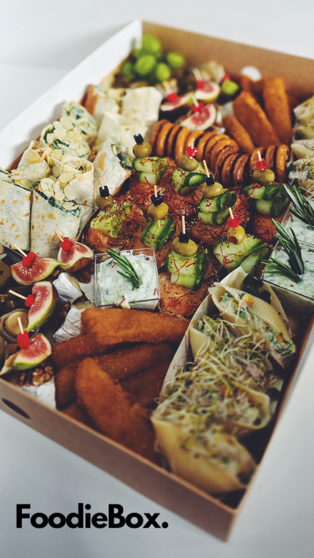 Elegancka kartonowa skrzynka z różnorodnymi przekąskami: roladki z łososiem, koreczki z ogórkiem i oliwką, tortille i inne smakołyki. Idealne na imprezę firmową.