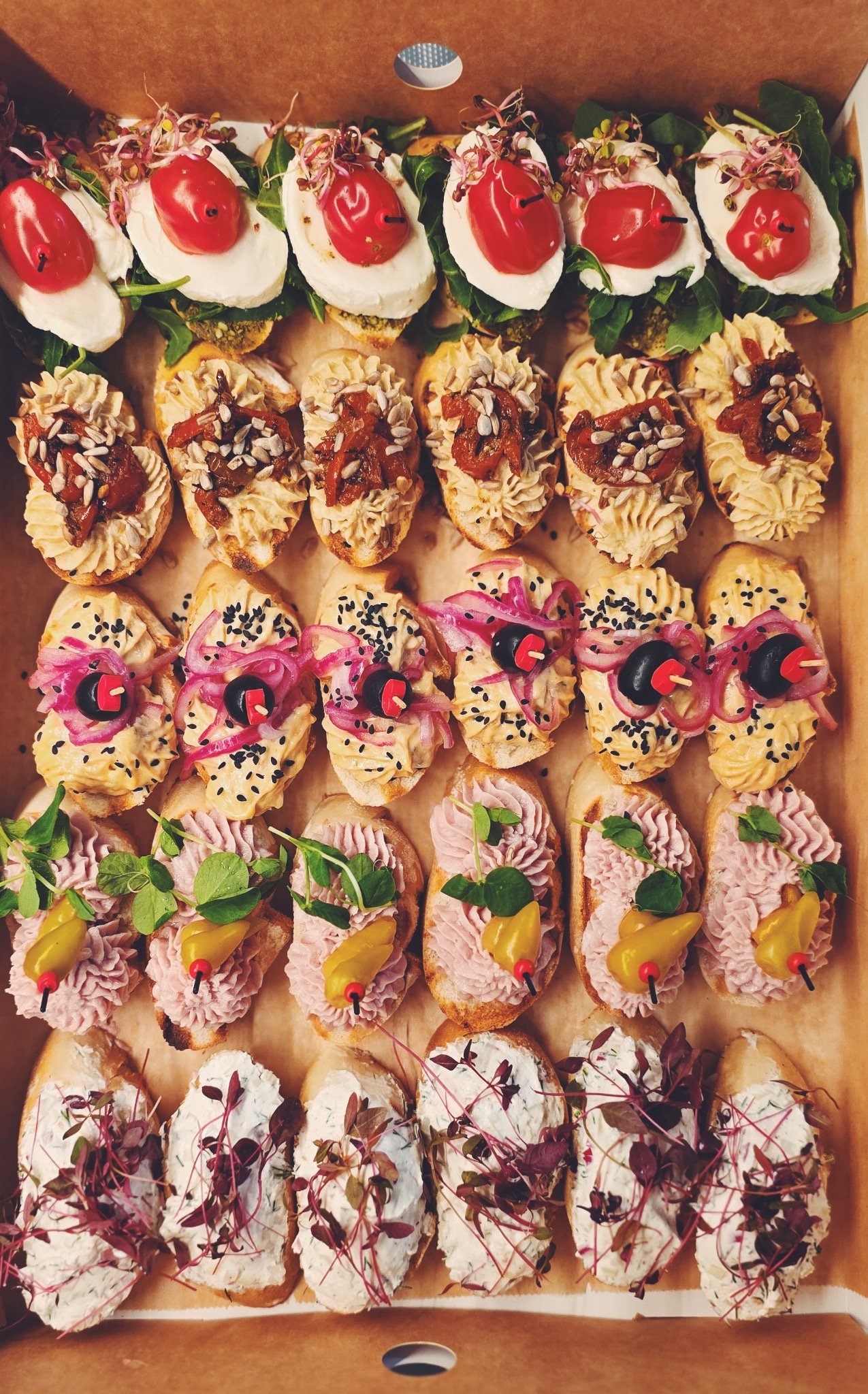 Różnorodne kanapki na pieczywie w kartonowym pudełku: z mozzarellą i pomidorem, pastą z suszonymi pomidorami, pastą z cebulą, różową pastą i białym serem.