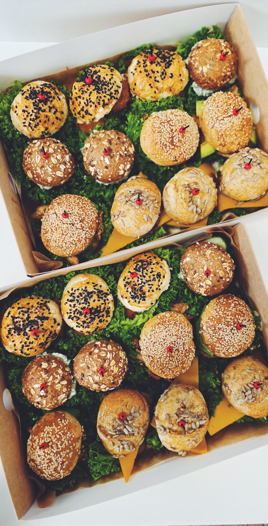 Dwa kartony pełne mini kanapek z różnymi posypkami (sezam, słonecznik, płatki owsiane), ułożonych na zielonej sałacie, z czerwonymi dekoracjami.