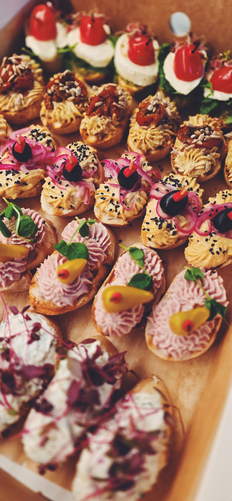 Różnorodne kanapeczki na jasnym pieczywie, z kremowymi pastami i kolorowymi dodatkami, ułożone w rzędach, gotowe do podania na przyjęciu.