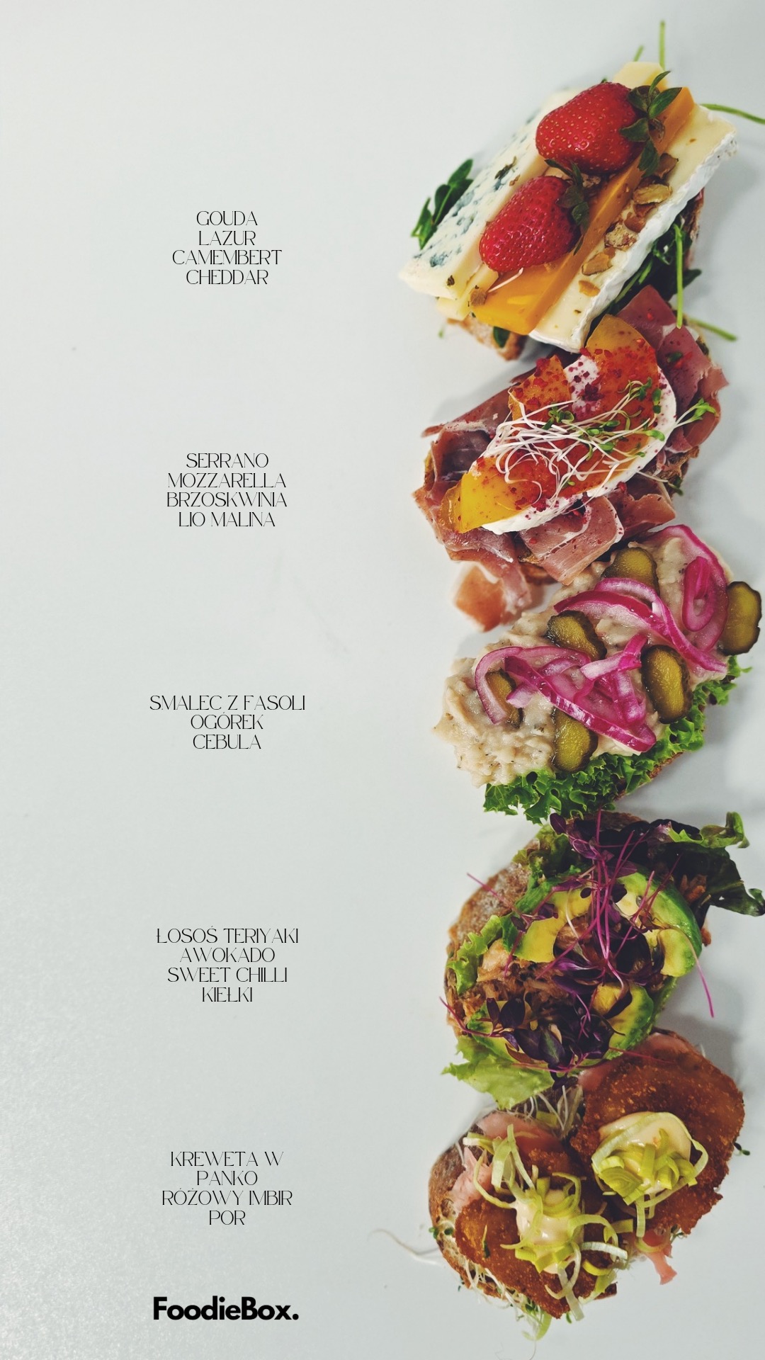 Kanapki z różnymi dodatkami: serami, wędlinami, warzywami i owocami, ułożone pionowo na białym tle. Widoczne nazwy składników. Elegancka kompozycja.