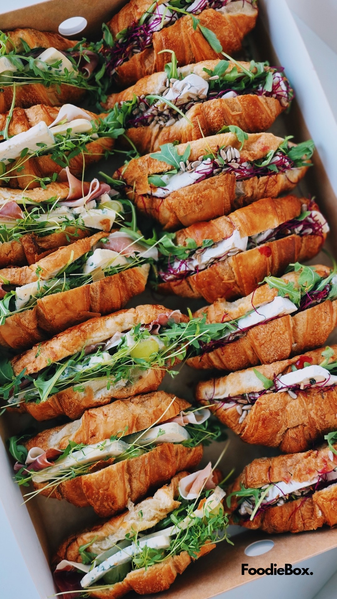 Karton z różnorodnymi croissantami: z szynką, serem pleśniowym, rukolą i kiełkami, idealne na catering. Widok z góry, estetyczne ułożenie.