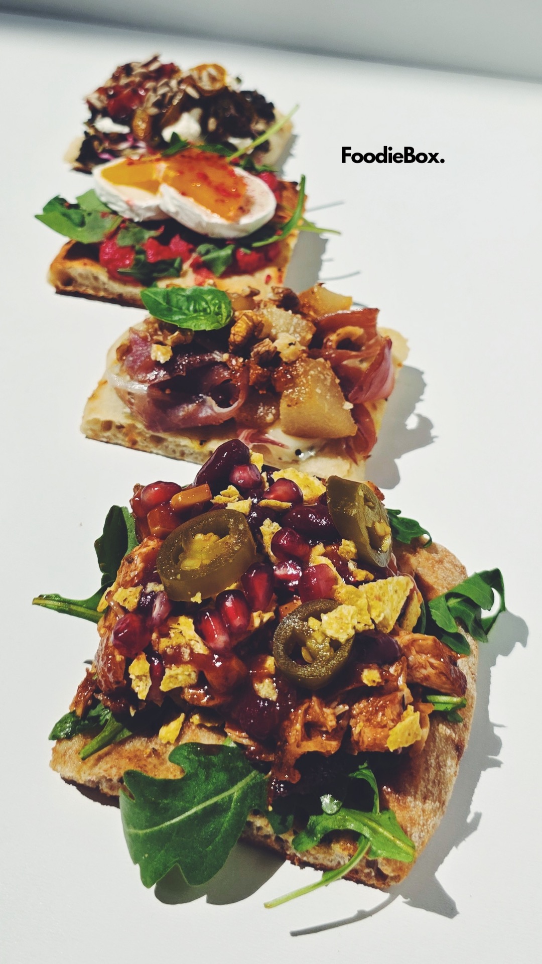 Cztery kanapki na jasnym tle, każda z unikalnymi składnikami: jalapeno, granat, ser kozi, karmelizowana cebula i orzechy. Widoczne logo 'FoodieBox.'.
