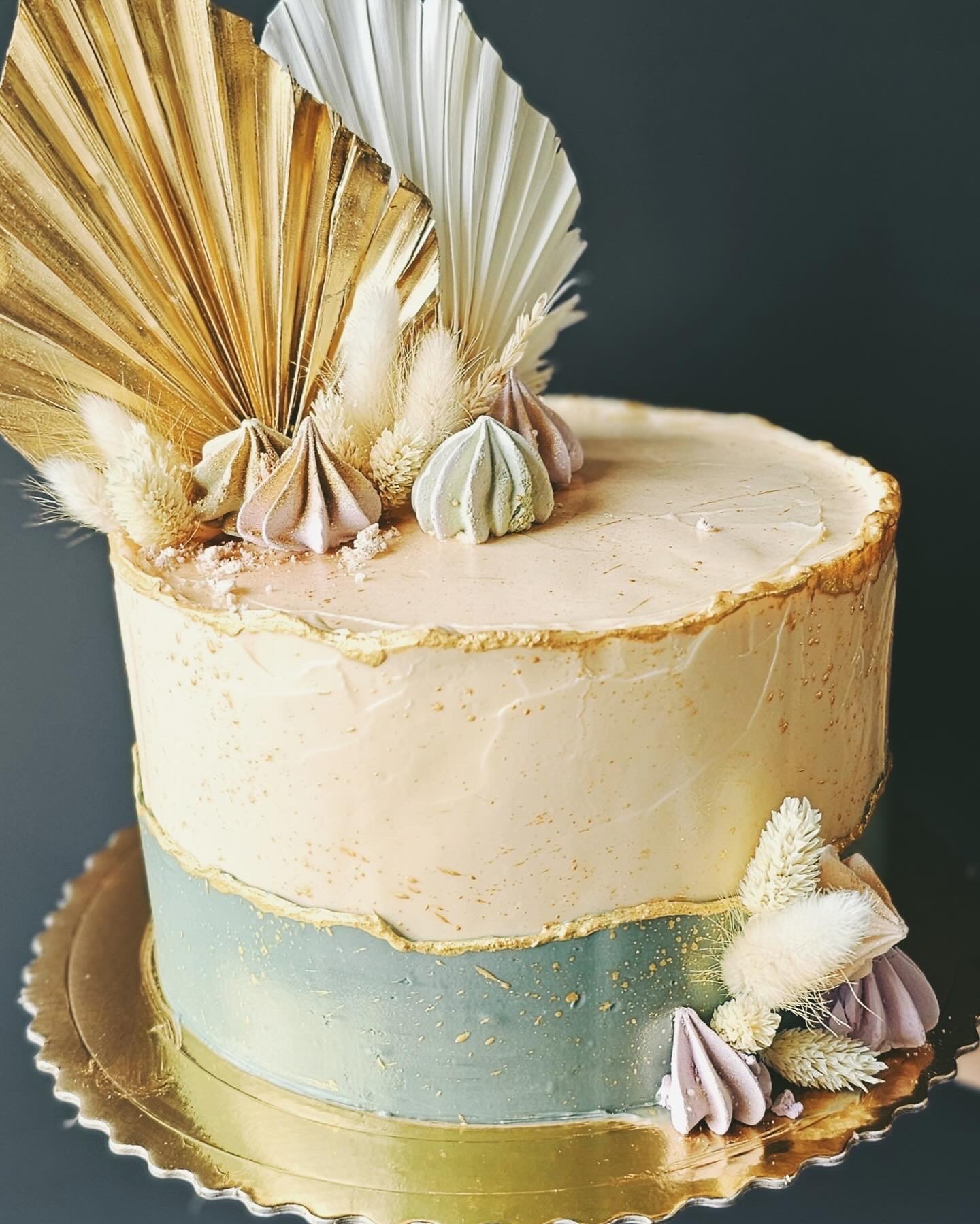 Elegancki tort z kremowym lukrem, złotymi akcentami i dekoracjami z suszonych traw oraz bezików na złotej paterze, na ciemnym tle.