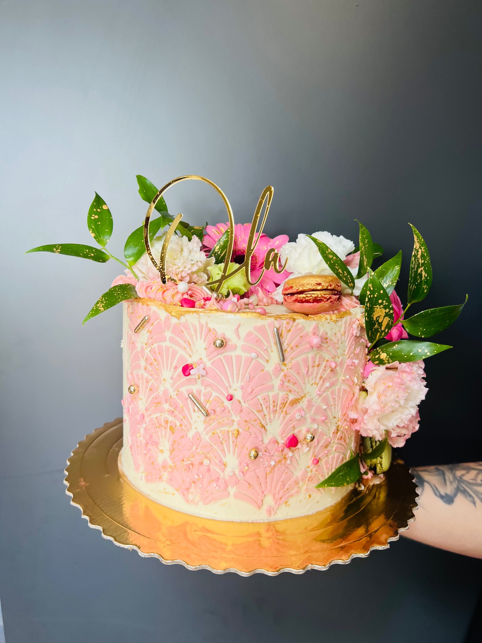 Ręcznie dekorowany tort w odcieniach różu i złota, zdobiony kwiatami, makaronikiem i napisem 'Ola', prezentowany na złotej paterze.