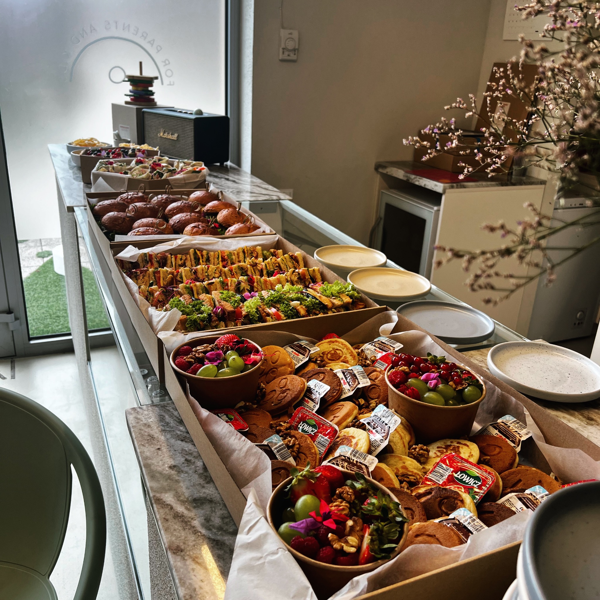 Stół cateringowy z różnorodnymi przekąskami: mini burgery, kanapki, owoce w miseczkach i ciasteczka. Widoczne elementy dekoracyjne i zastawa. Jasne, naturalne światło.