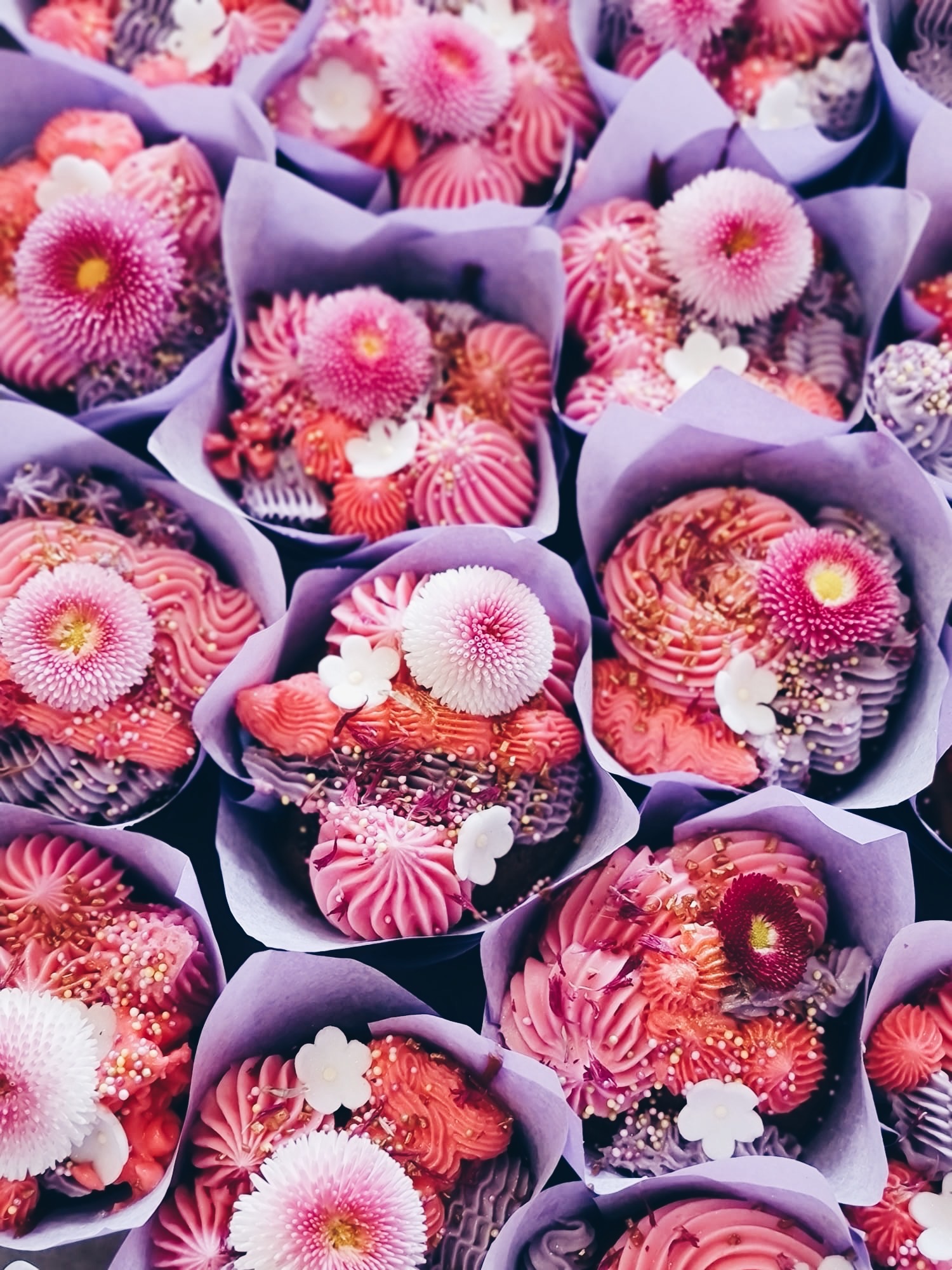 Różowe babeczki w liliowych papilotkach zdobione kremem, kwiatami i posypką, tworzące słodki bukiet. Widok z góry prezentuje bogactwo detali i artystyczne wykończenie.