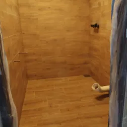 Wykończona kabina prysznicowa z brązowymi płytkami imitującymi drewno, widoczne elementy instalacji hydraulicznej i częściowo zakryte folią zabezpieczającą futryny drzwi.