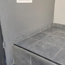 Granitowe płytki podłogowe i schodowe w odcieniach szarości z czarną listwą antypoślizgową na krawędzi stopnia, szara ściana z fakturą i biała skrzynka rewizyjna z okrągłym otworem.