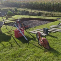 Przygotowanie podłoża pod kostkę brukową: wykop z podsypką żwirową, narzędzia Hilti, piła do betonu i poziomica na trawniku, w tle pole kukurydzy.