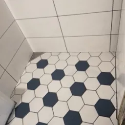 Podłoga w kabinie prysznicowej wyłożona biało-granatowymi heksagonalnymi płytkami imitującymi wzór piłki nożnej, białe płytki ścienne z czarnymi fugami, odpływ liniowy w rogu.