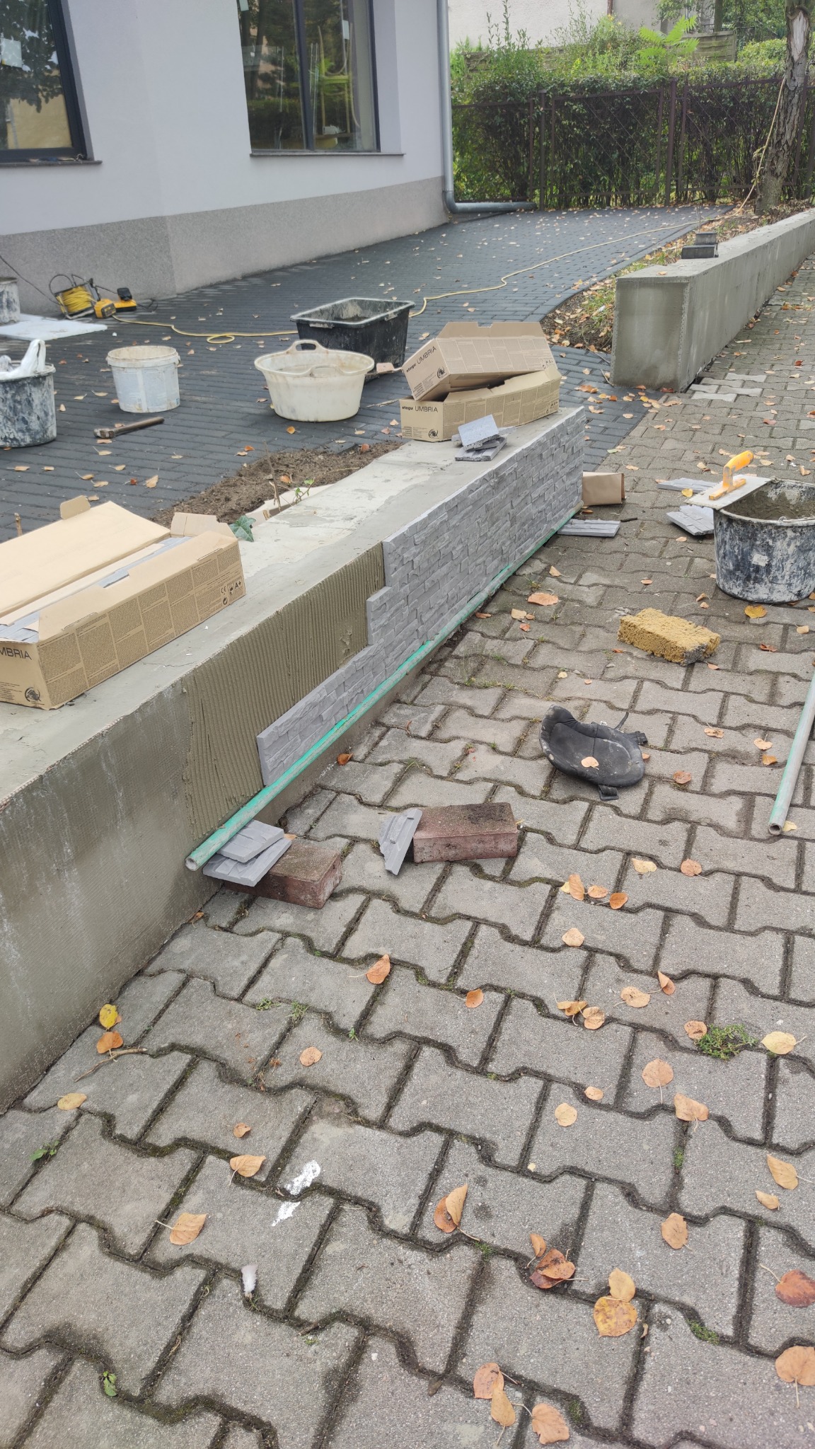 Montaż okładziny imitującej kamień na betonowym murku, widoczne paczki z okładziną, narzędzia i kostka brukowa na podłożu, jesienne liście.