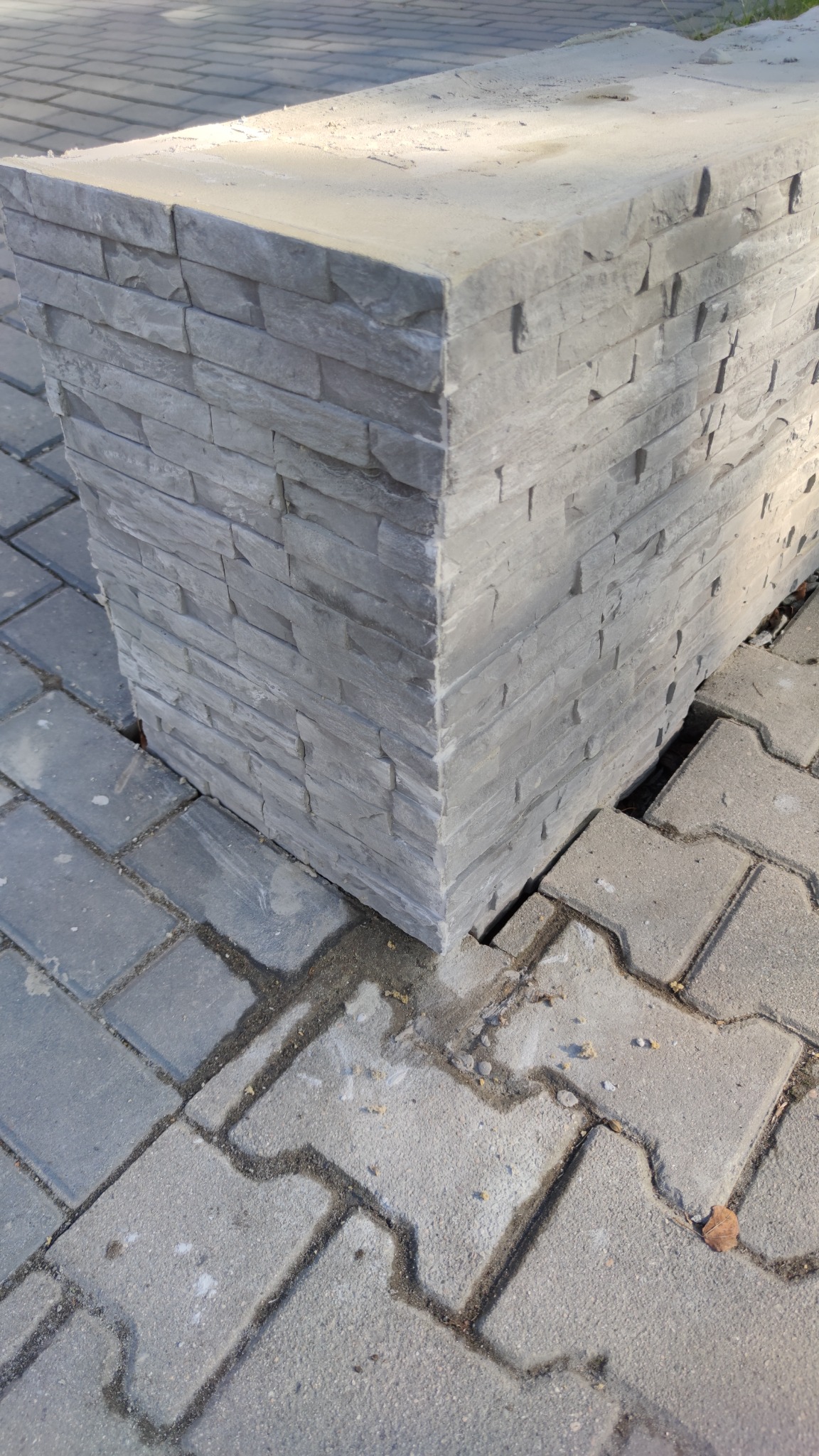 Szary, betonowy słupek z imitacją kamienia na kostce brukowej, widoczne niedokładności w dopasowaniu kostki wokół słupka.