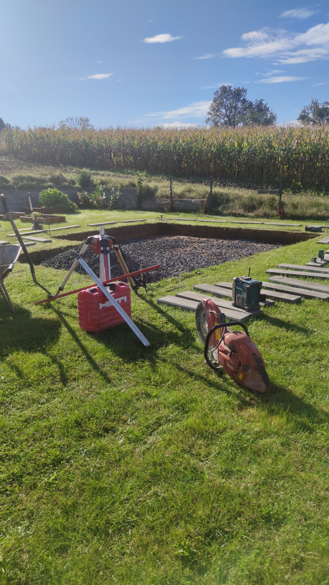 Przygotowanie podłoża pod kostkę brukową: wykop z podsypką żwirową, narzędzia Hilti, piła do betonu i poziomica na trawniku, w tle pole kukurydzy.
