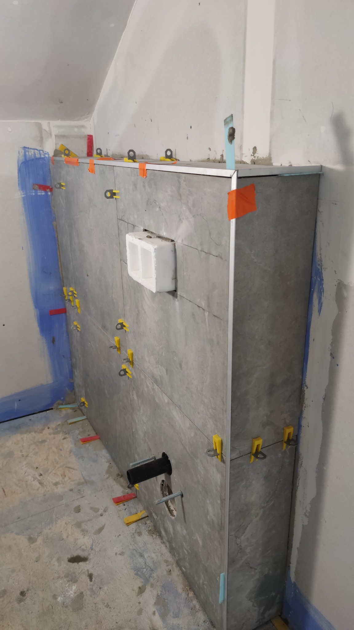 Ściana w trakcie układania płytek imitujących marmur, z widocznymi elementami poziomowania, otworem na instalację hydrauliczną i fragmentem niezabezpieczonej ściany z niebieską farbą.