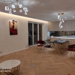 Stol-Lux - Jasne wnętrze mieszkania z parkietem ułożonym w jodełkę, ozdobnymi lampami sufitowymi, obrazem na ścianie i częściowo widoczną zabudową kuchenną.