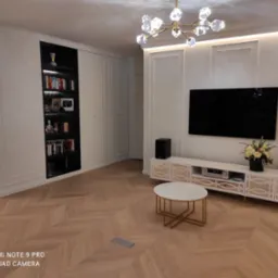 Elegancki salon z parkietem ułożonym w jodełkę, białe ściany z dekoracyjnymi panelami, szafa wnękowa z podświetlanymi półkami, telewizor oraz biała szafka RTV z dekoracyjnymi elementami i okrągły...