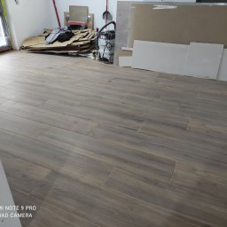 Stol-Lux - Ułożona podłoga z płytek imitujących drewno w trakcie prac wykończeniowych, widoczne kartony, odkurzacz przemysłowy, płyty gipsowo-kartonowe i resztki materiałów budowlanych.