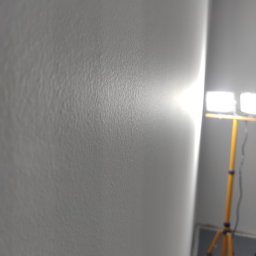 Stol-Lux - Świeżo wyszpachlowana ściana oświetlona lampą roboczą na statywie, widoczne drobne nierówności faktury.
